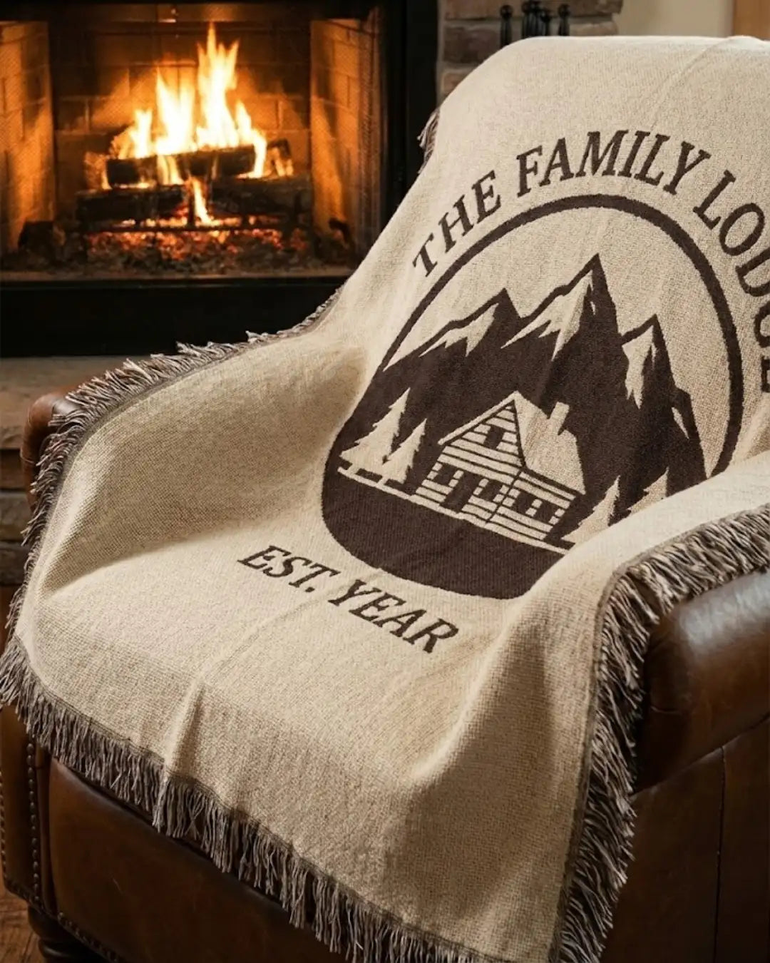 Beige woven blanket with black embroidered mountain cabin design and fringed edges.