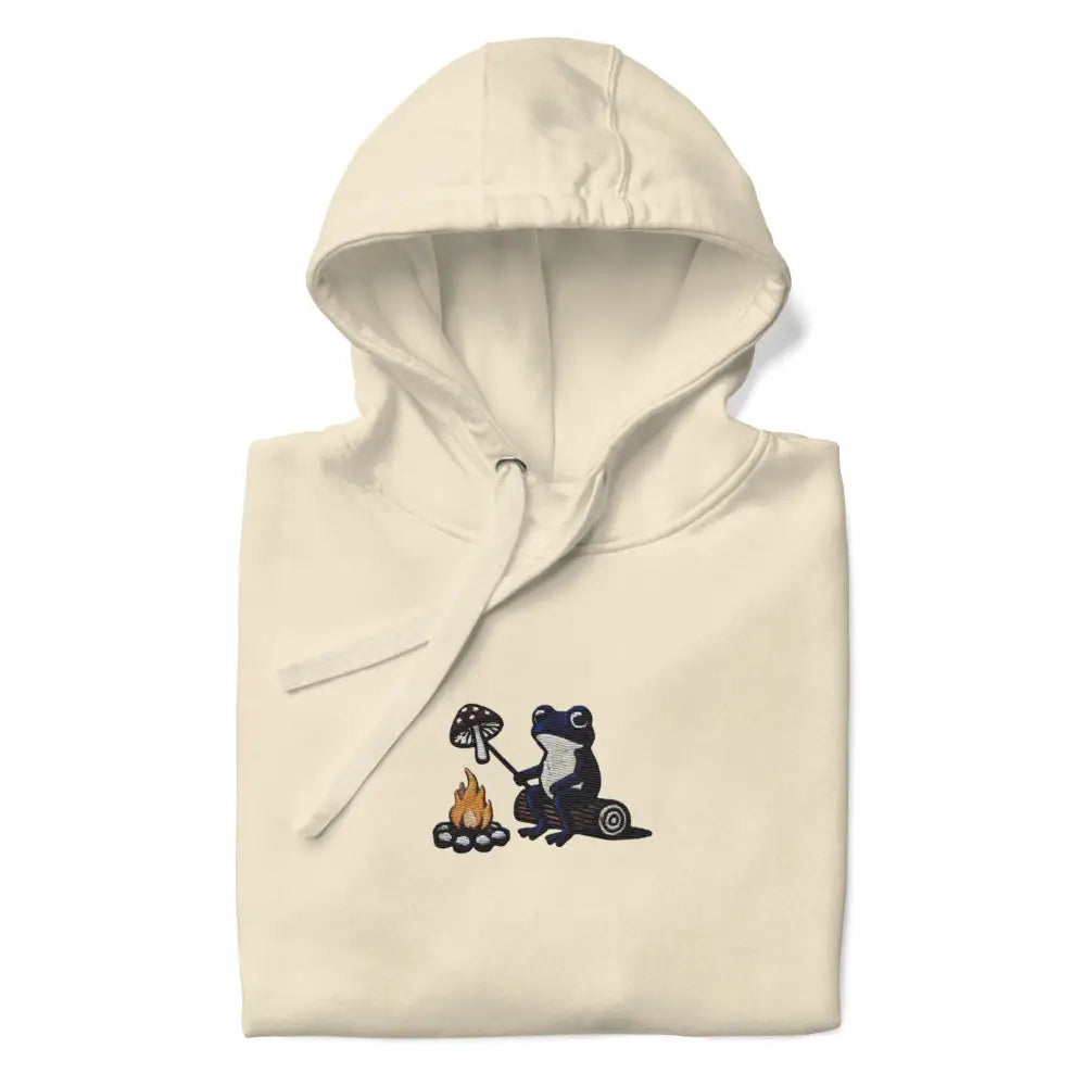 Cream-colored unisex hoodie with embroidered frog roasting mushroom over campfire